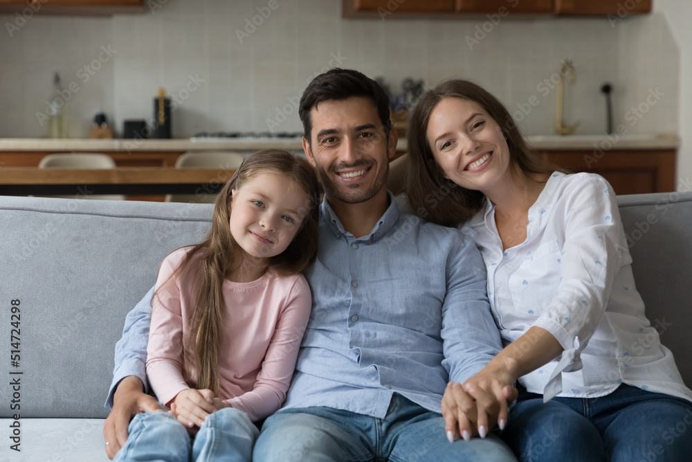 Beautiful multi ethnic family portrait, bank loan, tenancy concept. Preschool daughter, her young lovely mother and handsome Hispanic stepfather smile staring at camera posing seated on sofa at home Beautiful multi ethnic family portrait, bank loan, tenancy concept. Preschool daughter, her young lovely mother and handsome Hispanic stepfather smile staring at camera posing seated on sofa at home