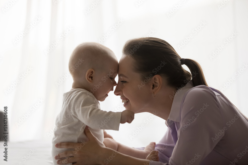 Happy motherhood, tender moments of mother and 0-6 baby, babyhood concept. Beautiful infant seated on bed touch foreheads with young loving mom feeling love, express caress, enjoy affection, close up Happy motherhood, tender moments of mother and 0-6 baby, babyhood concept. Beautiful infant seated on bed touch foreheads with young loving mom feeling love, express caress, enjoy affection, close up