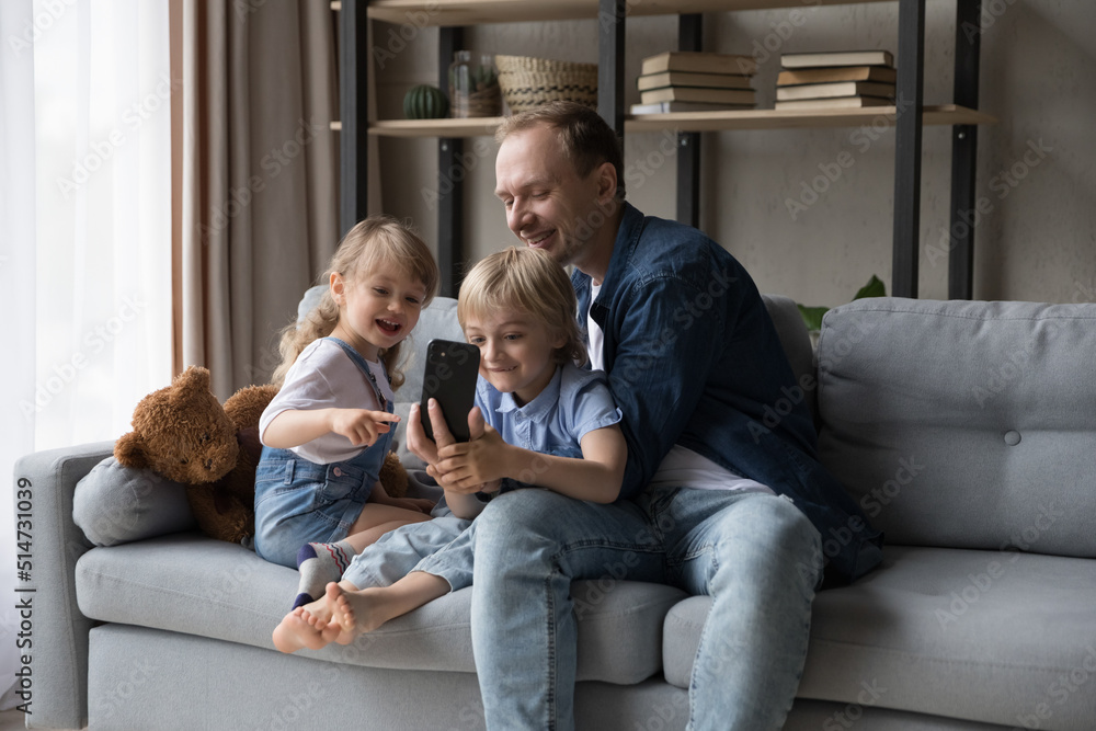 Loving dad and siblings enjoy new mobile application sit on sofa at home. Preschool girl and by spend time on internet under caring father control, watch new cartoons or video vlog. Fun, tech concept
