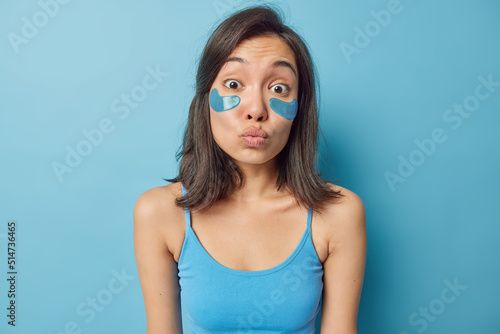 Lovely brunette young Asian woman applies beauty patches under eyes for skin treatment keepslips rounded dressed in t shirt isolated over blue background. Daily care and spa procedures concept