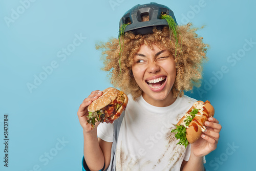 Cheerful woman has glad expression eats delicious fast food enjoys tasty snack winks eye smiles happily wears protective helmet and dirty t shirt isolated over blue background empty space for promo