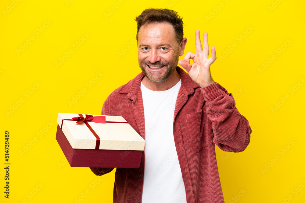 Middle age caucasian man holding a gift isolated on yellow background showing ok sign with fingers Middle age caucasian man holding a gift isolated on yellow background showing ok sign with fingers