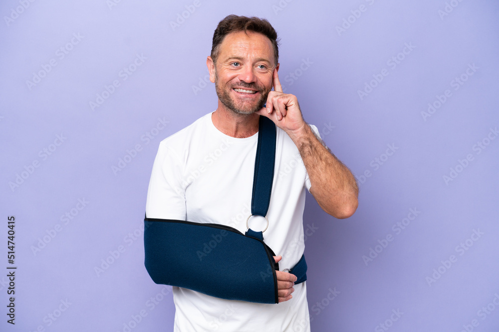 Middle age caucasian man with broken arm and wearing a sling isolated on purple background thinking an idea while looking up