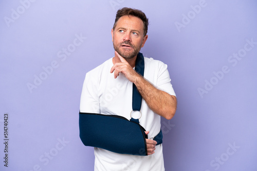 Middle age caucasian man with broken arm and wearing a sling isolated on purple background looking up while smiling