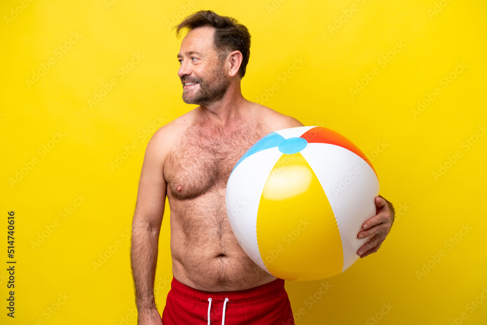 Middle age caucasian man holding beach ball isolated on yellow background looking side