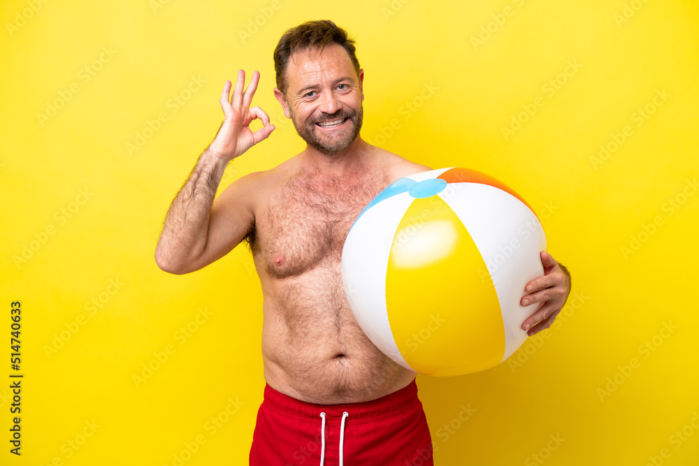 Middle age caucasian man holding beach ball isolated on yellow background showing ok sign with fingers