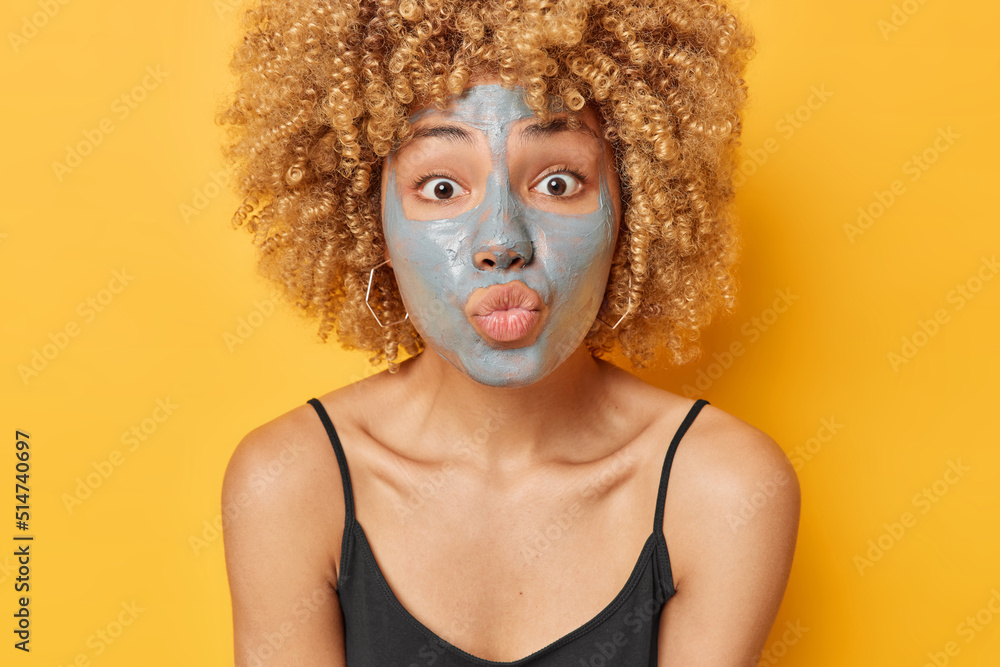 Indoor shot of beautiful woman with facial clay mask to treat acne and get more youthful looking skin keeps lips folded dressed in black t shirt isolated over yellow background. Daily routines