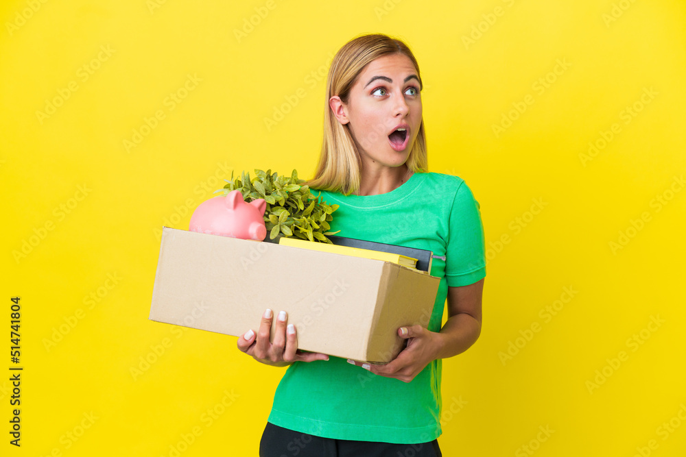 Young Uruguayan girl making a move while picking up a box full of things isolated on yellow background looking up and with surprised expression Young Uruguayan girl making a move while picking up a box full of things isolated on yellow background looking up and with surprised expression