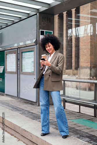 young latin woman waiting at the tram stop checking her mobile phone, concept of urban and modern lifestyle