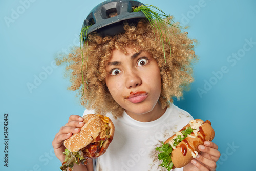 Indoor shot of shocked woman has curly hair holds tasty hamburger and hot dog eats unhealthily suffers from addiction to fast food wears protective helmet white dirty t shirt isolated on blue wall photo