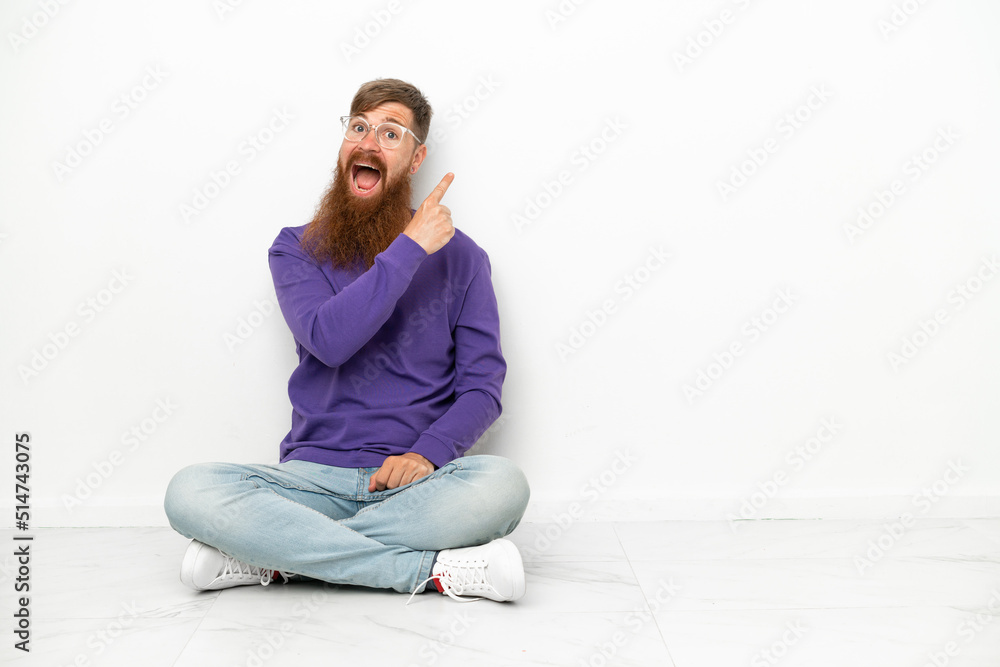Young caucasian reddish man sitting on the floor isolated on white background surprised and pointing side Young caucasian reddish man sitting on the floor isolated on white background surprised and pointing side