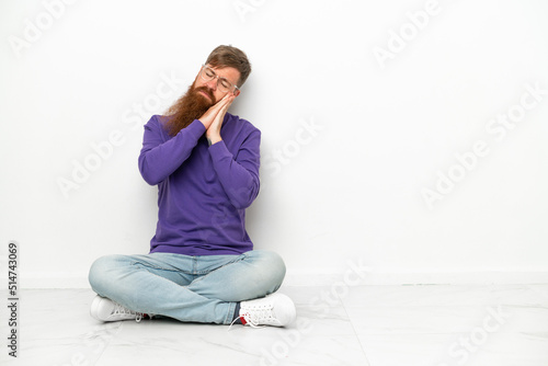 Young caucasian reddish man sitting on the floor isolated on white background making sleep gesture in dorable expression