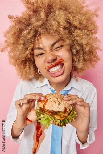 Vertical shot of cheerful curly haired woman eats delicious burger smeared with ketchup winks eyes clenches teeth wears white formal shirt with tie isolated over pink background. Unhealthy eating