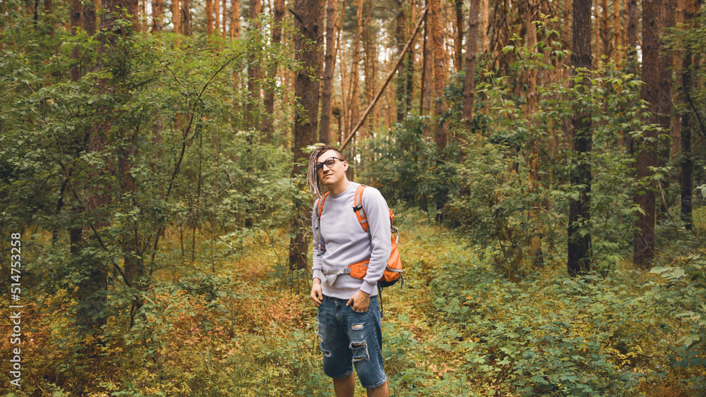 Obraz premium A man with a backpack poses in the woods. A male traveler with dreadlocks standing in a summer forest with a backpack