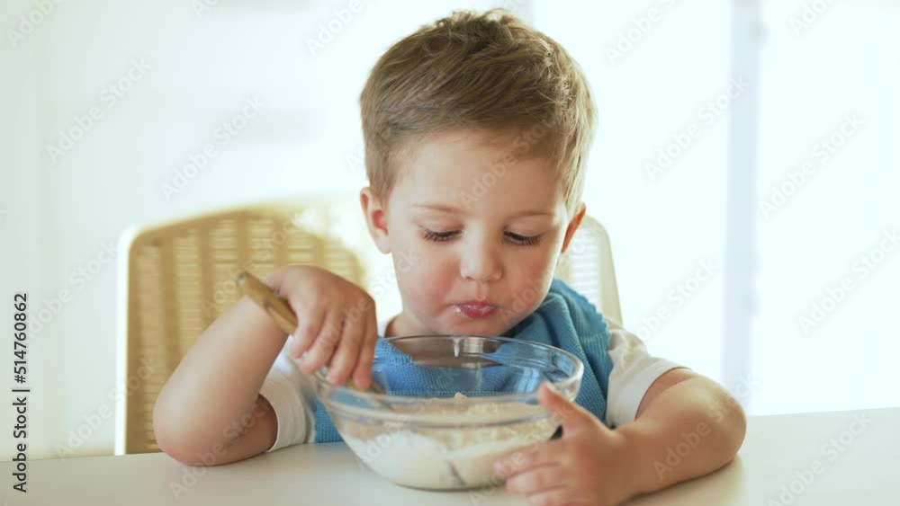 Baby eats cereal porridge. Bboy is holding spoon in his hand. Child sits at table and eats porridge.Family breakfast in kitchen. Feeding child at table.Child proper nutrition.Breakfast in kindergarten Baby eats cereal porridge. Bboy is holding spoon in his hand. Child sits at table and eats porridge.Family breakfast in kitchen. Feeding child at table.Child proper nutrition.Breakfast in kindergarten