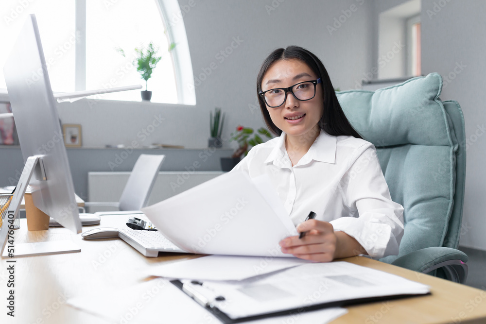 Portrait of young successful Asian financier, businesswoman working in office with documents, smiling and looking at camera. Portrait of young successful Asian financier, businesswoman working in office with documents, smiling and looking at camera.
