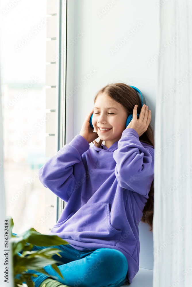 Cute little girl with wireless headphones sitting near window. Child listening favourite music in mobile application and smiling. Leisure at home Cute little girl with wireless headphones sitting near window. Child listening favourite music in mobile application and smiling. Leisure at home