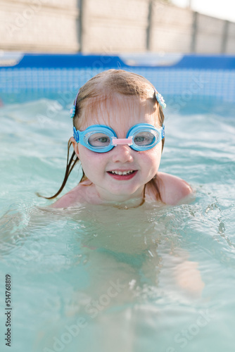 Little baby in swimming goggles, girl swim in paddling pool. Diving kid. Learning child to swim. Summer leisure and family holidays and vacation concept. People swim in a metal frame pool in backyard.