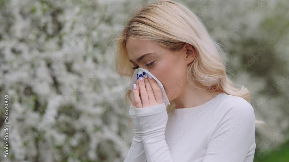 Young woman hiding her face with white handkerchief while sneezing outdoors. Caucasian lady with running nose standing near spring trees. Concept of seasonal illness. Young woman hiding her face with white handkerchief while sneezing outdoors. Caucasian lady with running nose standing near spring trees. Concept of seasonal illness.