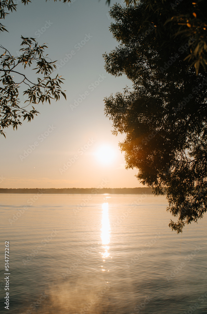 Sunrise on lake in morning, vertical landscape. Bright sun and light fog on water in sunny summer