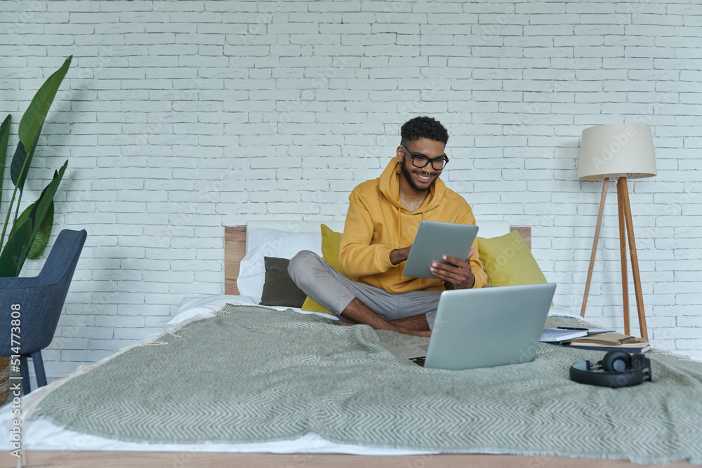 Cheerful African man using technologies while sitting on the bed at home