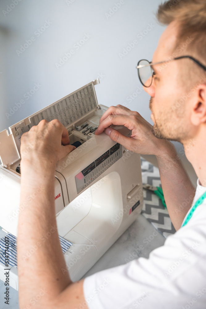 Obraz premium A blond man with glasses changes a bobbin in a sewing machine. A tailor with a measuring tape on his neck in a white t-shirt. The concept of small business, lifestyle and enjoying of work