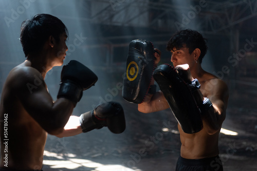Strong Asian sportsman practicing boxing workout punching with male combat sport trainer in abandoned building. Active man coach support male boxer do fighting exercise kickboxing in dark old gym © CandyRetriever 
