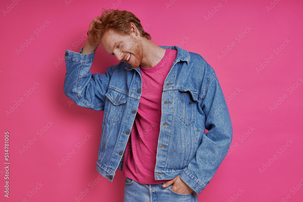 Handsome redhead man in denim clothing holding hand ion hair against pink background Handsome redhead man in denim clothing holding hand ion hair against pink background