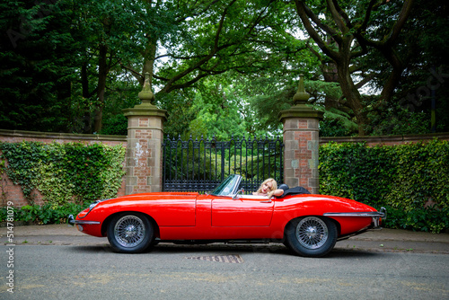 Lady with long blond hair resting in a red E-Type Jaguar with gates and park in the background