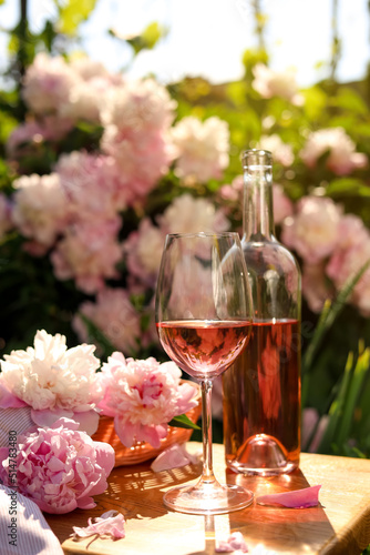 Bottle and glass of rose wine near beautiful peonies on wooden table in garden