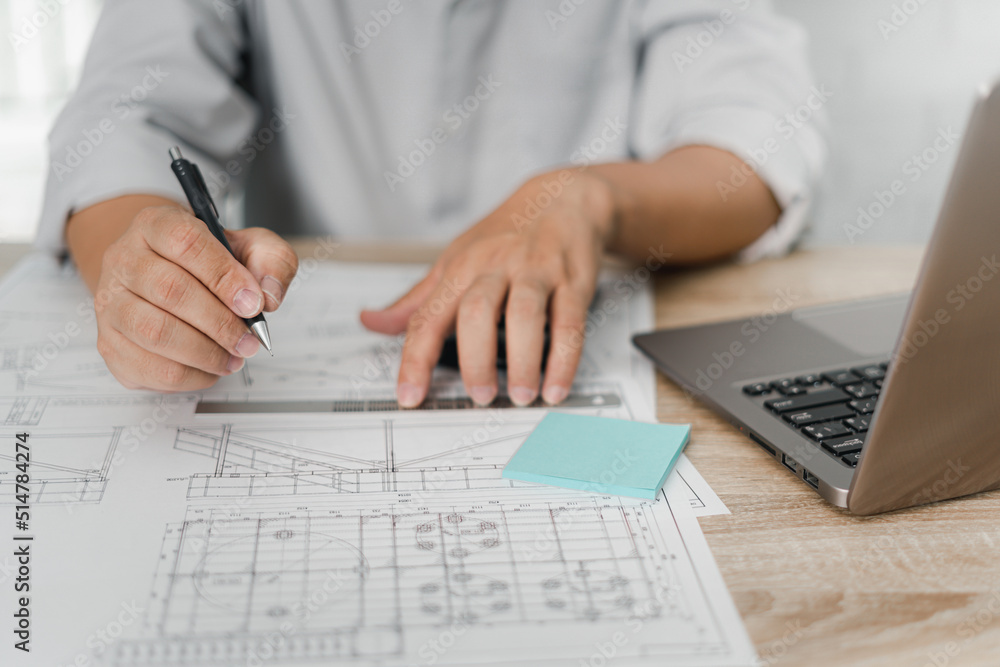 Fototapeta premium Engineering work concept: Close up hand a use pencil and steel ruler mark CAD drawing and calculator in Calculating document and laptop on the wood table in office