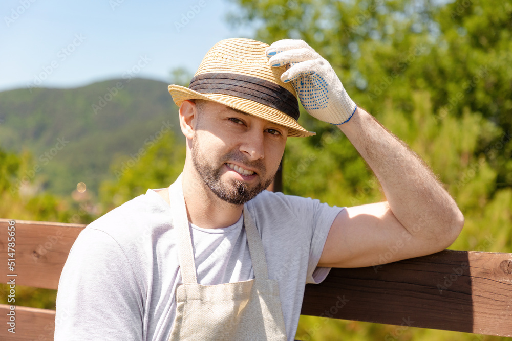 Portrait of a smiling handsome bearded gardener in gloves and apron wearing a straw hat, near a wooden fence. In the background is a sunny green yard. Gardening and farming
