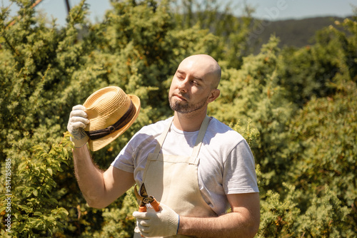 Sunstroke. Portrait of bald bearded man wearing in gloves and apron fanning with straw hat holding a plunger. In the background is a sunny garden trees. Summer heat and gardening