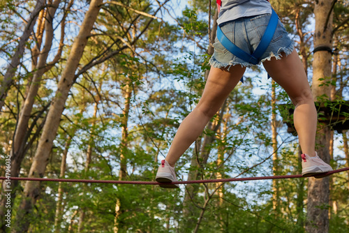 Young girl in denim shorts deftly walks a tightrope in a national nature park. Woman using safety equipment overcomes the track in a rope park against the background of huge pine trees