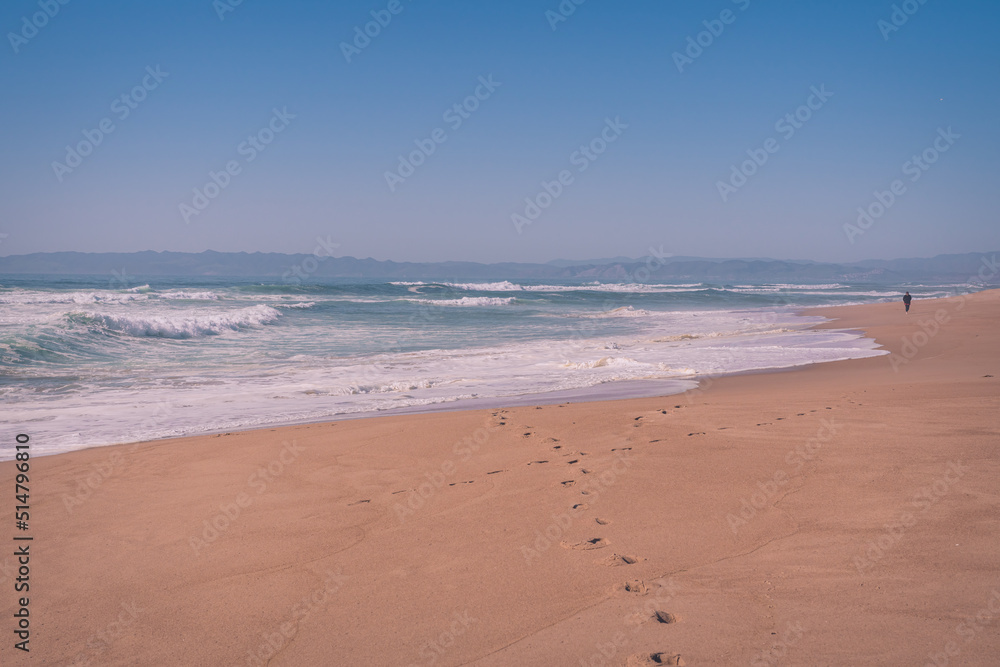Dreamlike seascape in light blue and pink colors. Ocean, wide sandy beach, foot prints on sand, and silhouette of walking man on a horizon