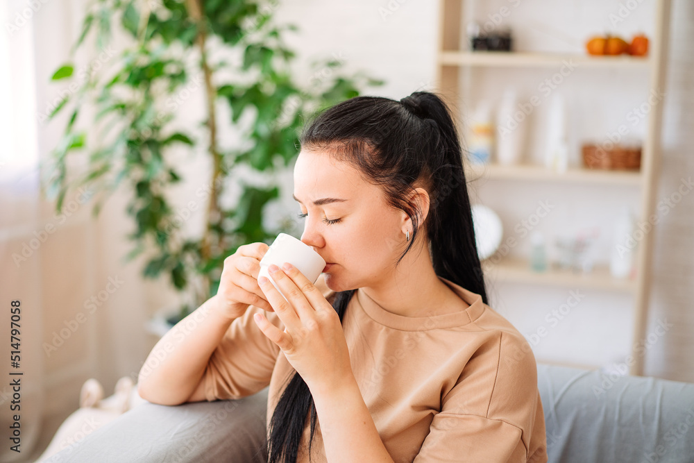 A happy young woman drinks a cup of tea on an autumn morning. A dreamy girl is sitting in the living room with a cup of hot coffee. A beautiful woman at home. A happy young woman drinks a cup of tea on an autumn morning. A dreamy girl is sitting in the living room with a cup of hot coffee. A beautiful woman at home.