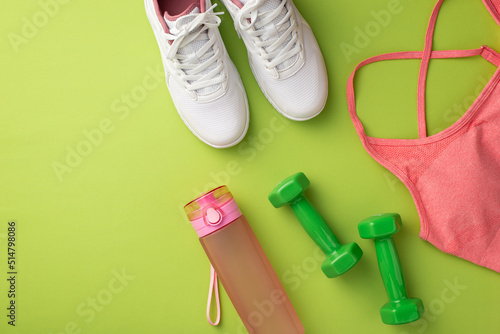 Fitness concept. Top view photo of pink sports bra sneakers bottle of water and green dumbbells on isolated green background