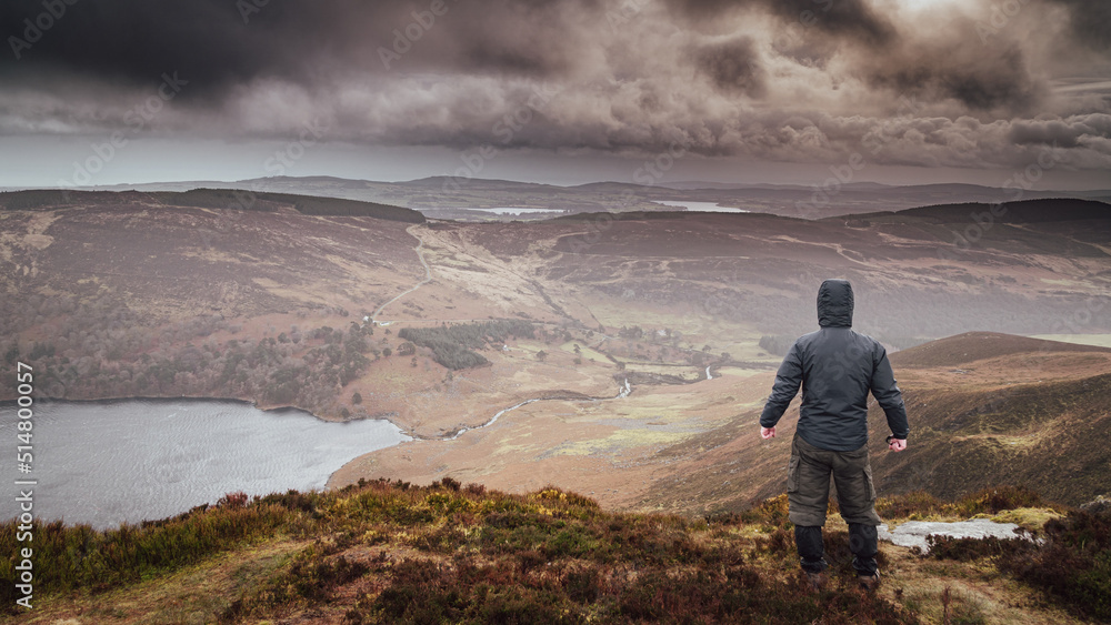 Man from behind looking towards Lough Tay (Guinness Lake) from Luggala peak in the Wicklow mountains Ireland. Cloudy and wet day