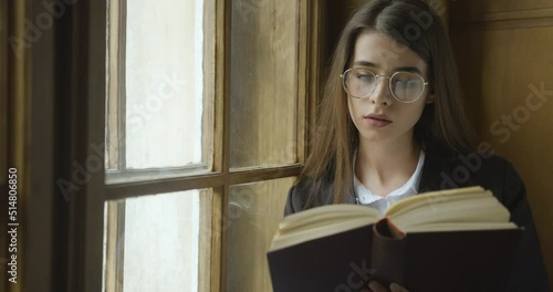 Young womat in glasses sitting on the windowsill and reading a book. Side view of a pretty woman resting on the windowsill near a large window. photo