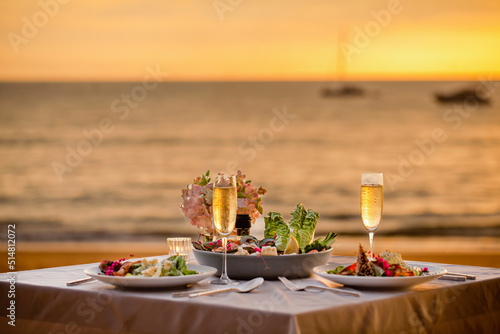 Romantic sunset dinner on beach. Table honeymoon set for two with luxurious food, glasses of champagne drinks in restaurant with sea view and yachts on background. Summer vacation or wedding concept. © TravelMedia