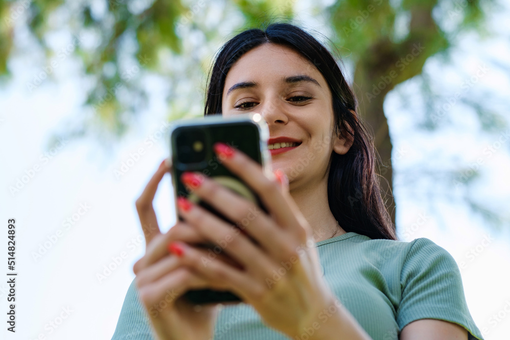 Happy woman wearing tee standing on city park, outdoors looking at the phone screen and using phone. Messaging with friends, watching video or scrolling on social media. Bottom up view. Happy woman wearing tee standing on city park, outdoors looking at the phone screen and using phone. Messaging with friends, watching video or scrolling on social media. Bottom up view.
