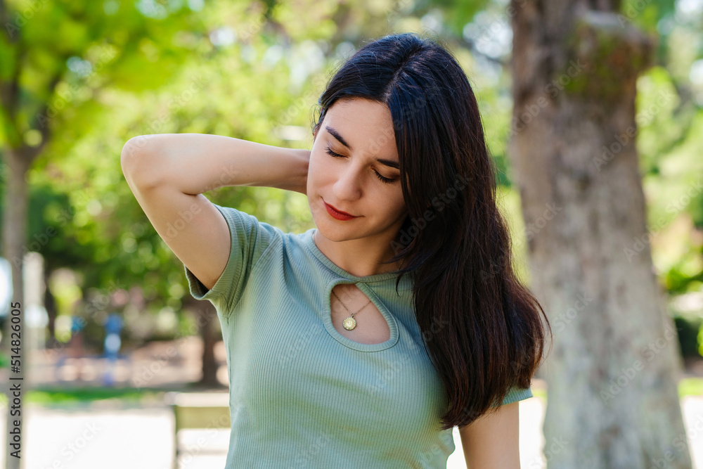 Beautiful brunette woman wearing turquoise t-shirt standing on city park, outdoors feeling hurt joint shoulder back pain ache, fibromyalgia concept, close up rear view. Health care.