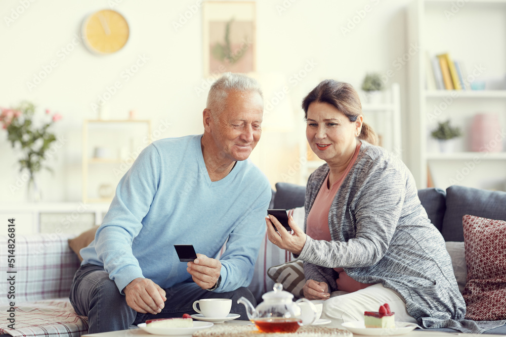 Positive elderly woman in cardigan sitting on sofa in living room and watching old photos with husband over tea cup Positive elderly woman in cardigan sitting on sofa in living room and watching old photos with husband over tea cup