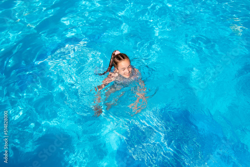 A teenage girl of 11-13 years old swims in a pool with blue water. She has african braids braided with zi-zi ribbons. Summer. Family vacation.