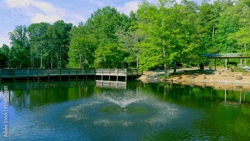 stunning aerial footage of a gorgeous summer landscape in the park with a lake and a water fountain and a brown wooden bridge surrounded by pink trees and lush green trees and grass with blue sky photo