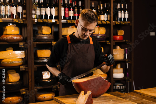 Handsome cheese sommelier with big knife for cut cheese. Bearded hipster in fashion glasses working in cheese shop. Creative idea for advertising.