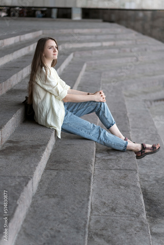 Portrait of girl sitting on the stairs. Modern young woman in jeans and shirt resting outside on the stairs.