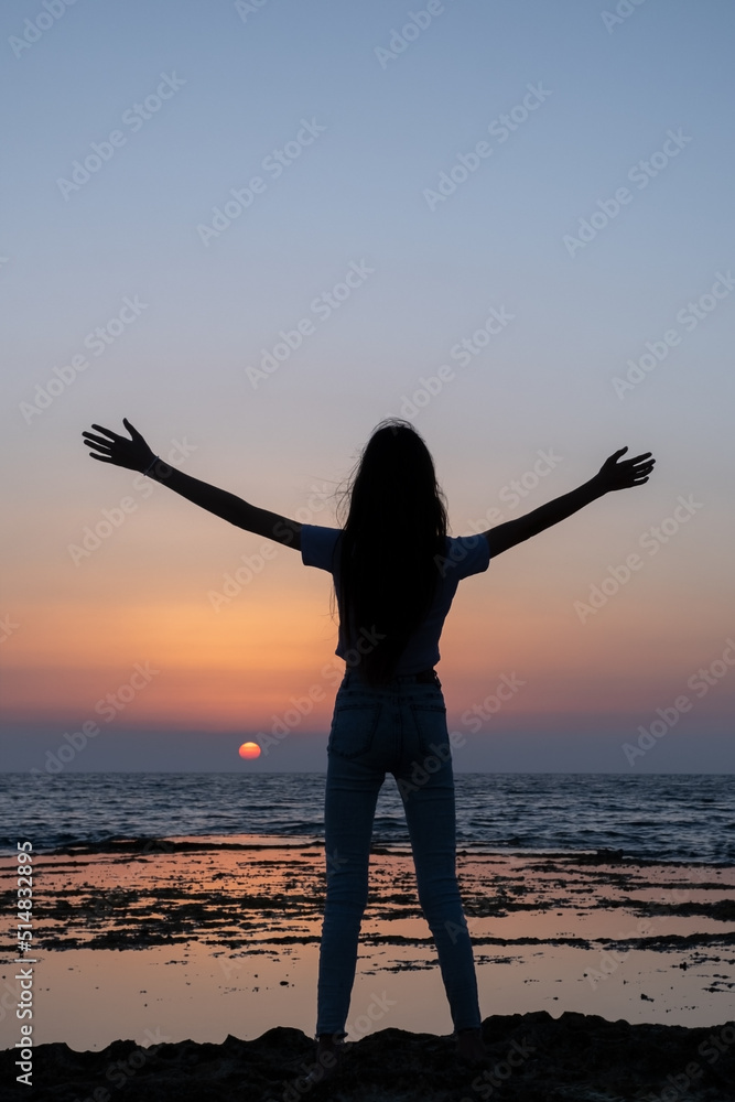 Teen woman looking to sunset, rejoicing by raising her hands up against the sea in israel. Triumph silhouette