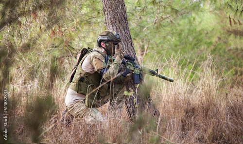 A military man with a machine gun in his hands gives a signal to another. An armed man near a tree. Combat strategy