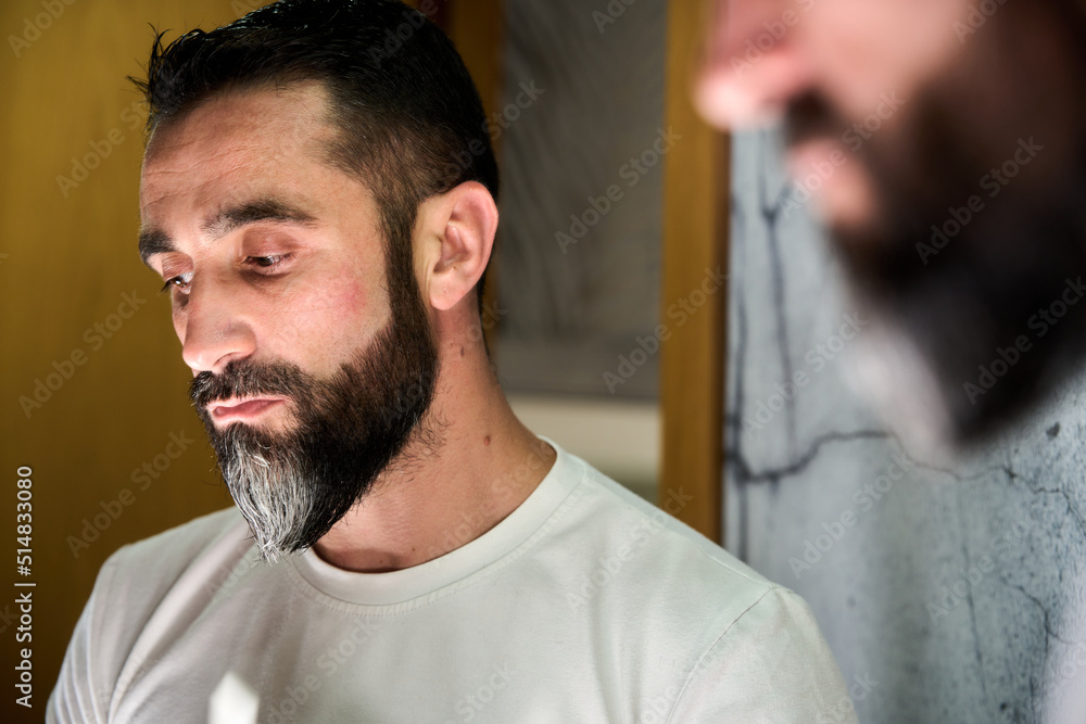 Fototapeta premium Young man grooming and combing his beard with comb in front of mirror
